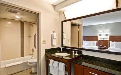 Modern hotel guest bathroom with a bathtub and shower, granite vanity with sink, large framed mirror, fresh white towels, and a view into a guest room with two beds.