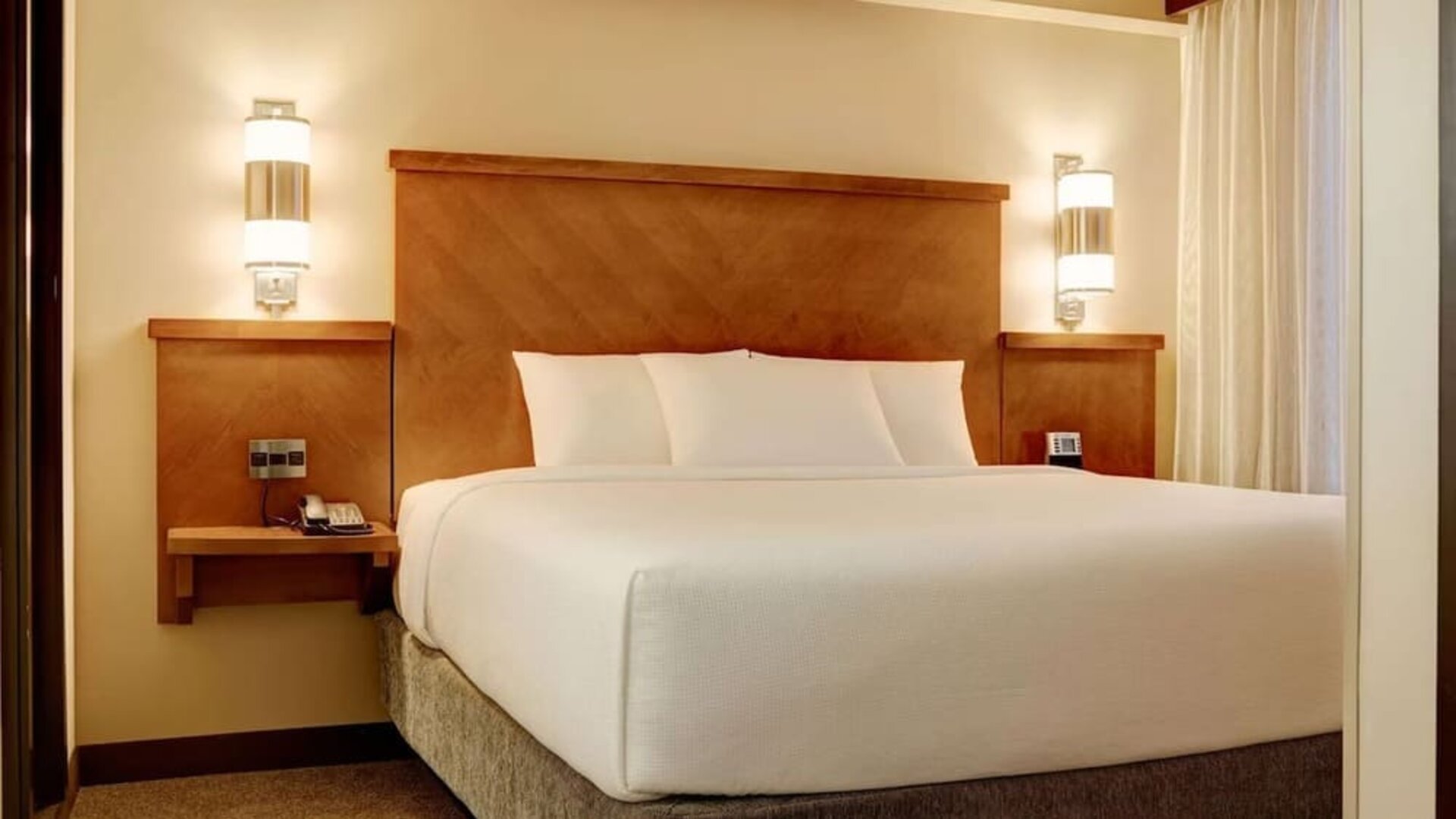 Comfortable hotel king bed dressed in crisp white linens and pillows, framed by a warm wooden headboard, bedside tables, contemporary wall lights, and a large window with curtains.