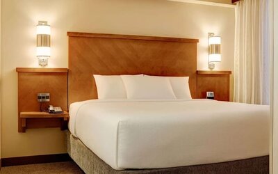 Comfortable hotel king bed dressed in crisp white linens and pillows, framed by a warm wooden headboard, bedside tables, contemporary wall lights, and a large window with curtains.