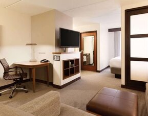 Spacious hotel room featuring a king bed with white linens, a separate sitting area with sectional sofa and ottoman, contemporary wood accents, wall lighting, and a partially visible TV.