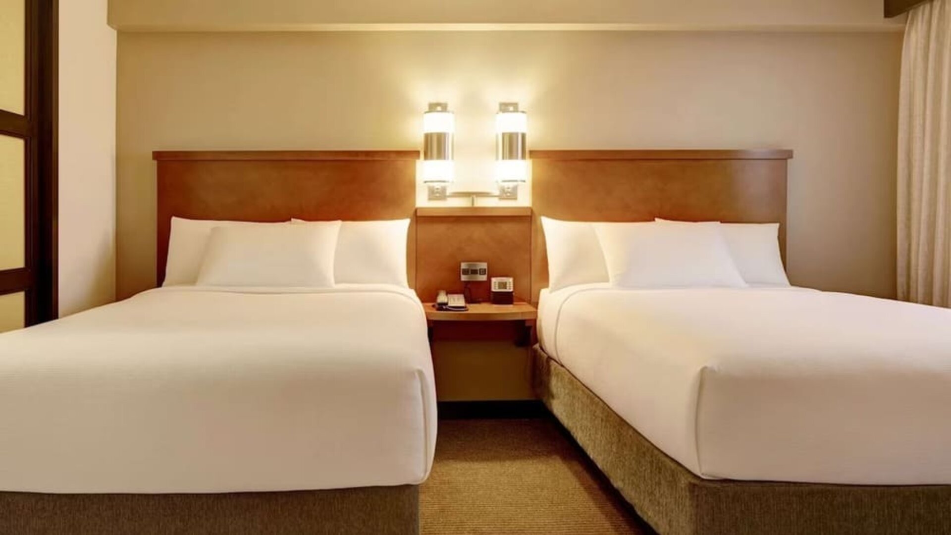 Bright hotel guest room with two beds dressed in crisp white linens, a shared wooden headboard and bedside table, modern wall lights, neutral carpeting, and warm beige décor.
