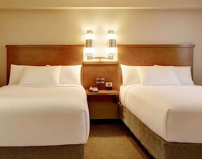 Bright hotel guest room with two beds dressed in crisp white linens, a shared wooden headboard and bedside table, modern wall lights, neutral carpeting, and warm beige décor.