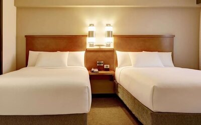 Bright hotel guest room with two beds dressed in crisp white linens, a shared wooden headboard and bedside table, modern wall lights, neutral carpeting, and warm beige décor.