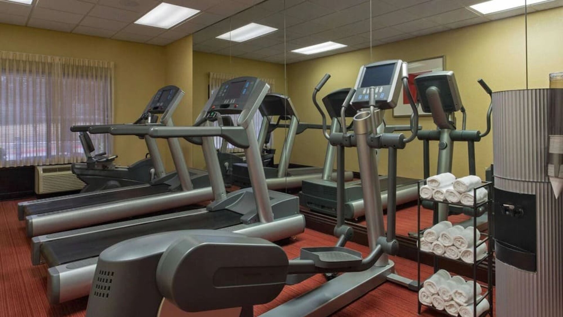 Hotel fitness center featuring treadmills, elliptical machines, cardio equipment, a mirrored wall, fresh towels, and large windows providing natural light.