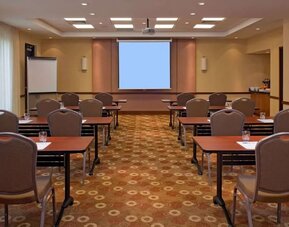 Hotel meeting and conference room arranged classroom-style with tables and chairs, a large projection screen, flip chart, ceiling-mounted projector, and refreshment station.