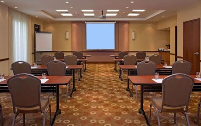 Hotel meeting and conference room arranged classroom-style with tables and chairs, a large projection screen, flip chart, ceiling-mounted projector, and refreshment station.