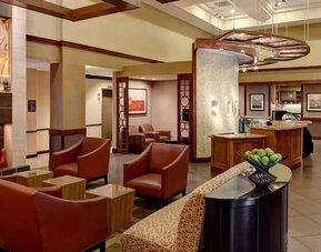 Stylish hotel lobby lounge with rust-colored armchairs, small side tables, contemporary artwork, warm wood accents, tiled flooring, and an open café area in the background.