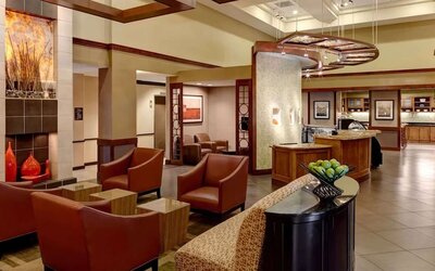 Stylish hotel lobby lounge with rust-colored armchairs, small side tables, contemporary artwork, warm wood accents, tiled flooring, and an open café area in the background.
