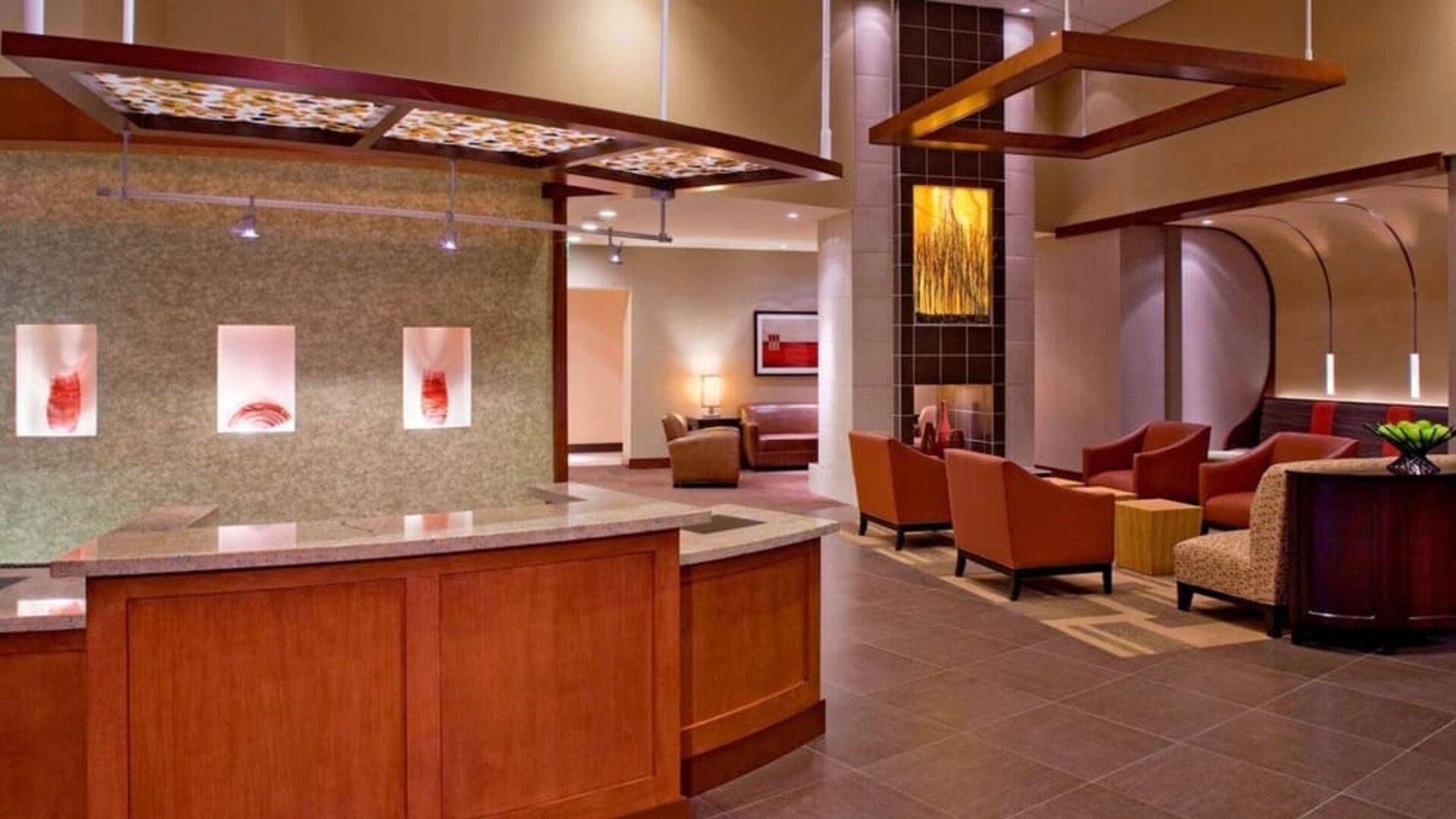 Contemporary hotel reception and lobby area featuring a wood and granite front desk, rust-colored lounge chairs, decorative wall art, modern overhead fixtures, and warm ambient lighting.