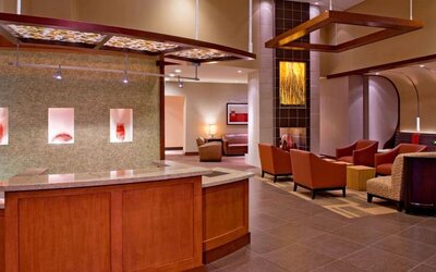 Contemporary hotel reception and lobby area featuring a wood and granite front desk, rust-colored lounge chairs, decorative wall art, modern overhead fixtures, and warm ambient lighting.