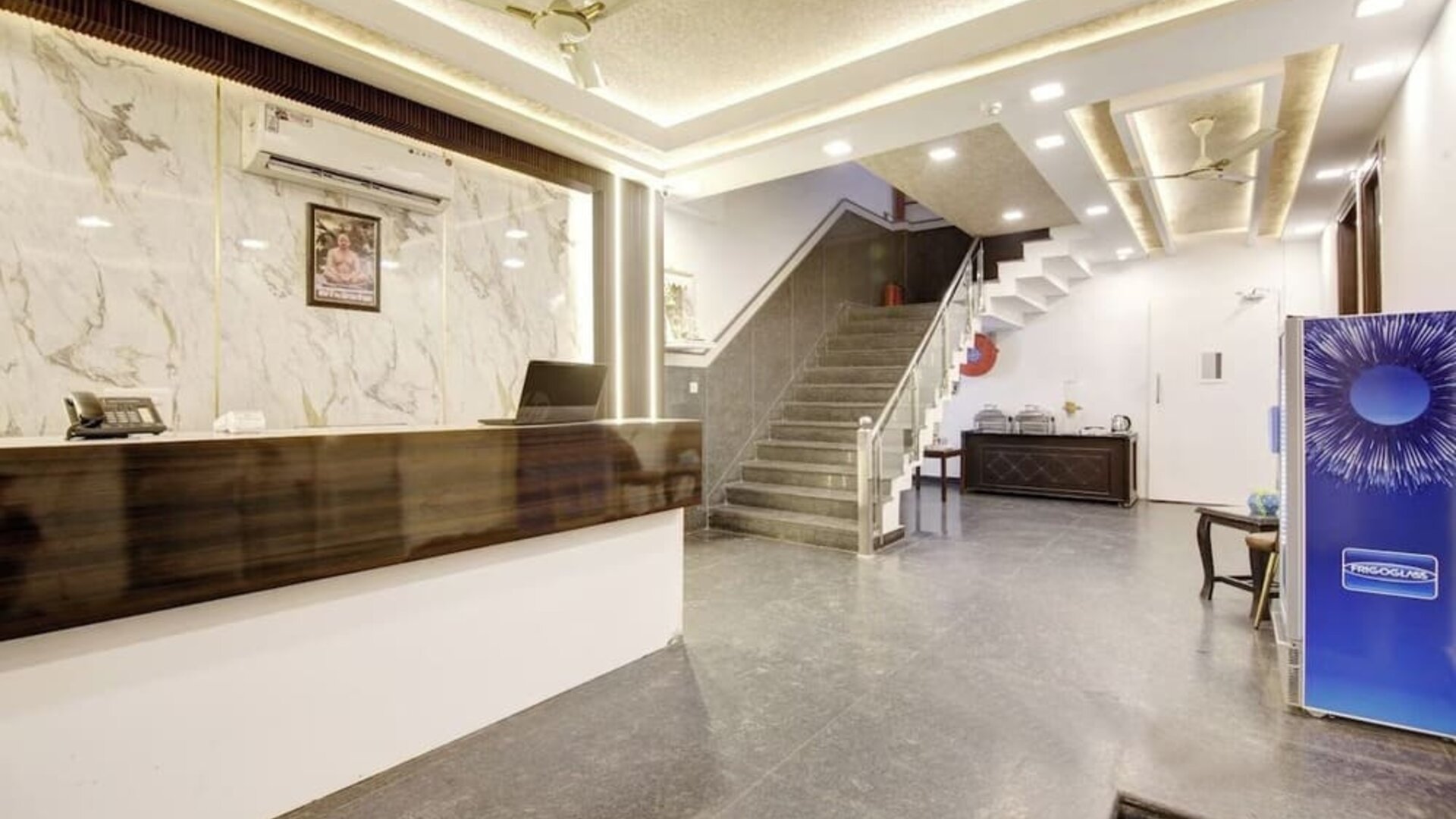 Spacious hotel lobby with a dark wood reception counter, marble-patterned wall, illuminated ceiling, gray tiled staircase with glass railing, seating area and beverage cooler.