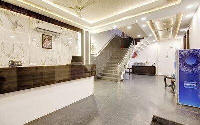 Spacious hotel lobby with a dark wood reception counter, marble-patterned wall, illuminated ceiling, gray tiled staircase with glass railing, seating area and beverage cooler.