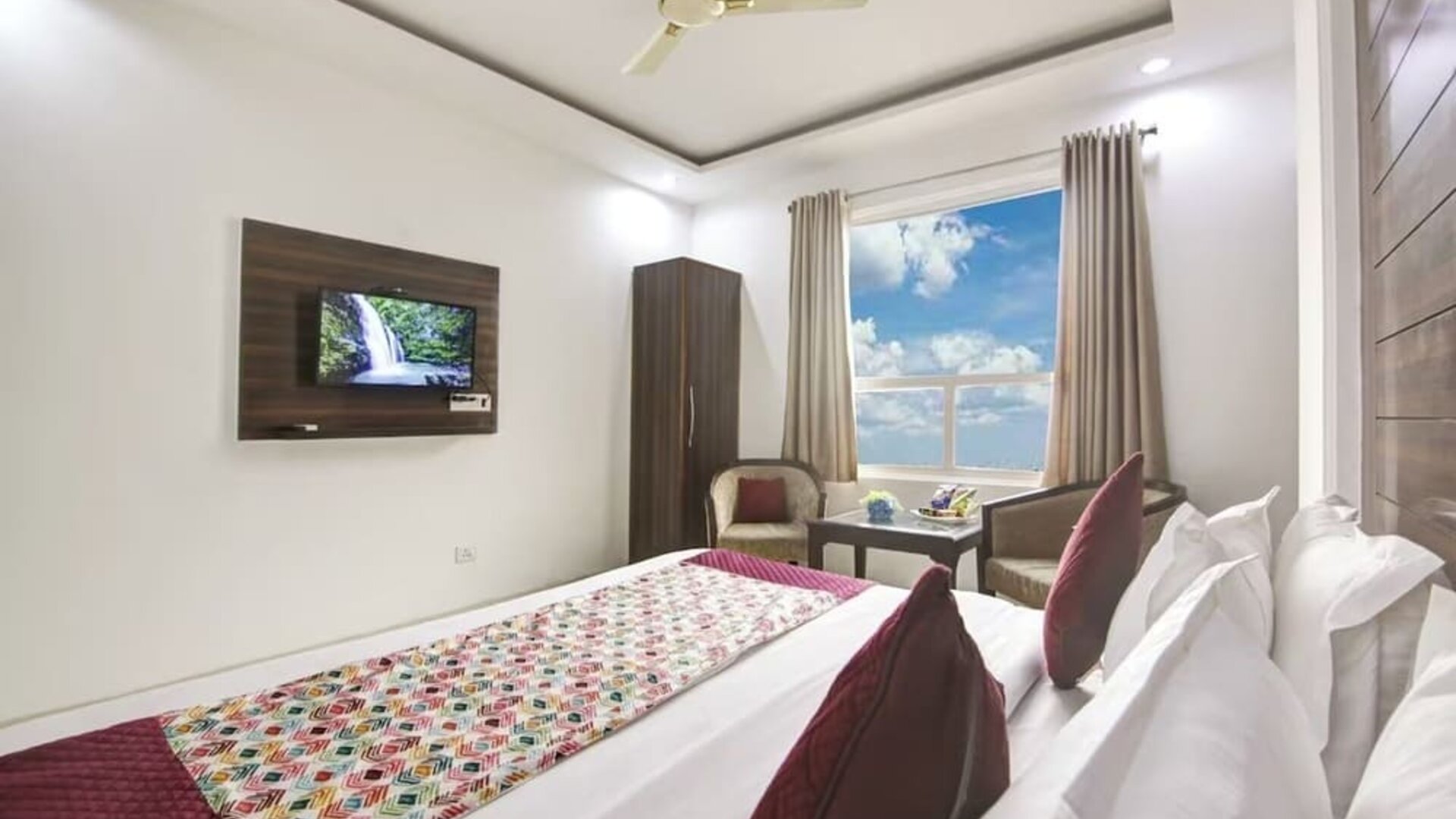 Modern hotel room with a large bed dressed in white linens and red patterned accents, dark wood feature headboard, ceiling fan, large window with curtains, wall-mounted TV, seating area and small glass-topped table.