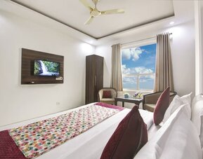 Modern hotel room with a large bed dressed in white linens and red patterned accents, dark wood feature headboard, ceiling fan, large window with curtains, wall-mounted TV, seating area and small glass-topped table.