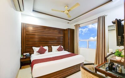 Bright hotel room with a large bed dressed in white linens, burgundy pillows and a colorful patterned runner, complemented by a dark wood headboard, wardrobe, two armchairs, small table and large window with curtains.