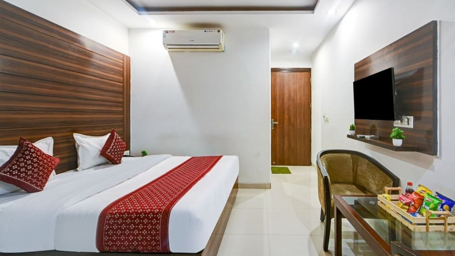 Contemporary hotel room with a large bed featuring white linens and red patterned accents, dark wood wall panels, wall-mounted TV, air conditioning, upholstered seating and a glass table with refreshments.