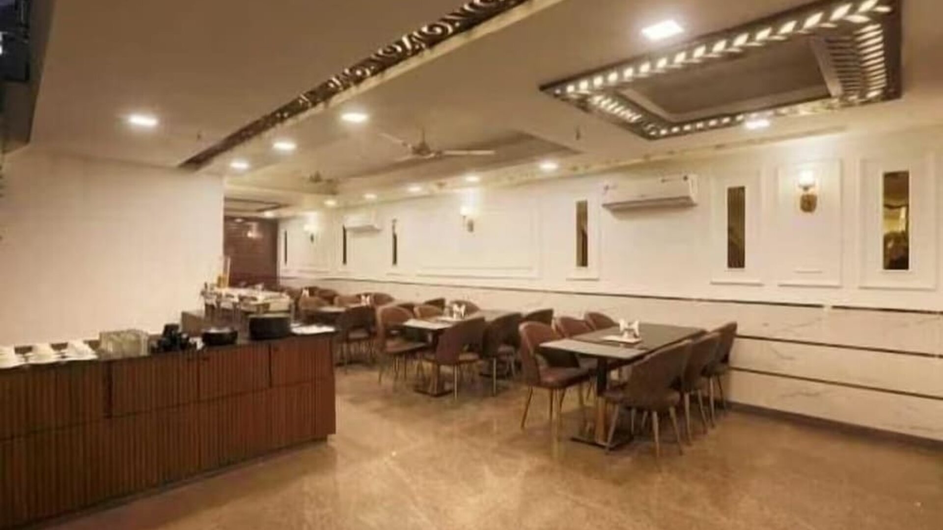 Spacious hotel dining area with multiple tables and upholstered chairs, buffet counter, polished flooring, decorative ceiling panels, recessed lighting, ceiling fans and wall-mounted air-conditioning units.