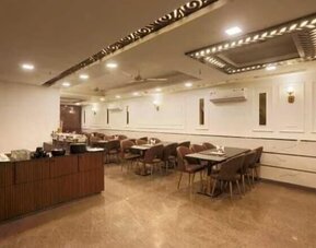 Spacious hotel dining area with multiple tables and upholstered chairs, buffet counter, polished flooring, decorative ceiling panels, recessed lighting, ceiling fans and wall-mounted air-conditioning units.