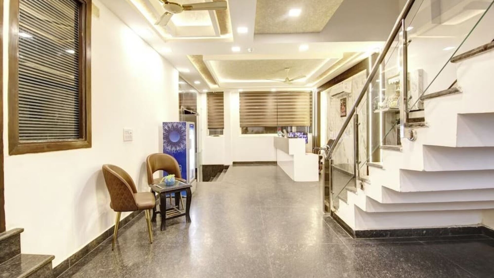 Bright interior hotel entrance and lobby corridor with polished stone flooring, two chairs and a small table, illuminated ceiling details, glass-railed staircase and reception area visible in the background.