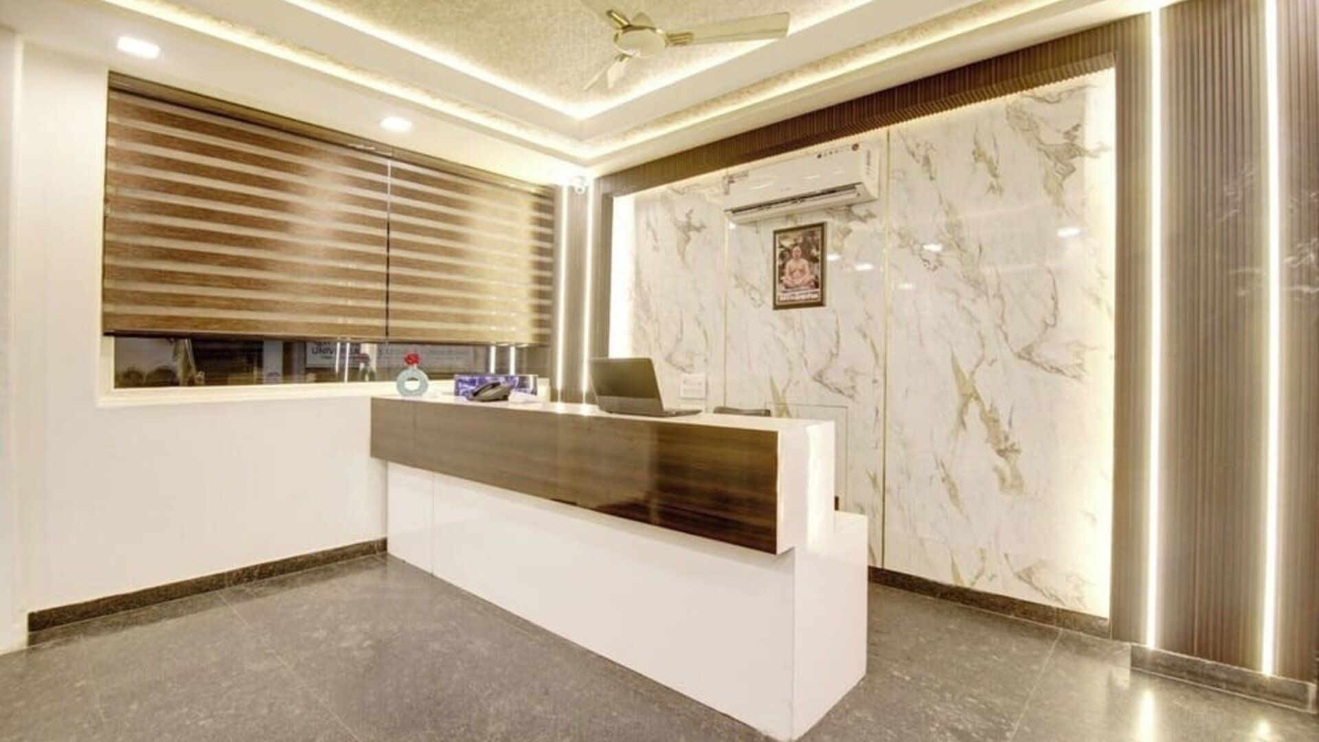 Modern hotel reception desk with dark wood and white finishes, marble-patterned feature wall, vertical accent lighting, large window with striped blinds, ceiling fan and polished gray flooring.