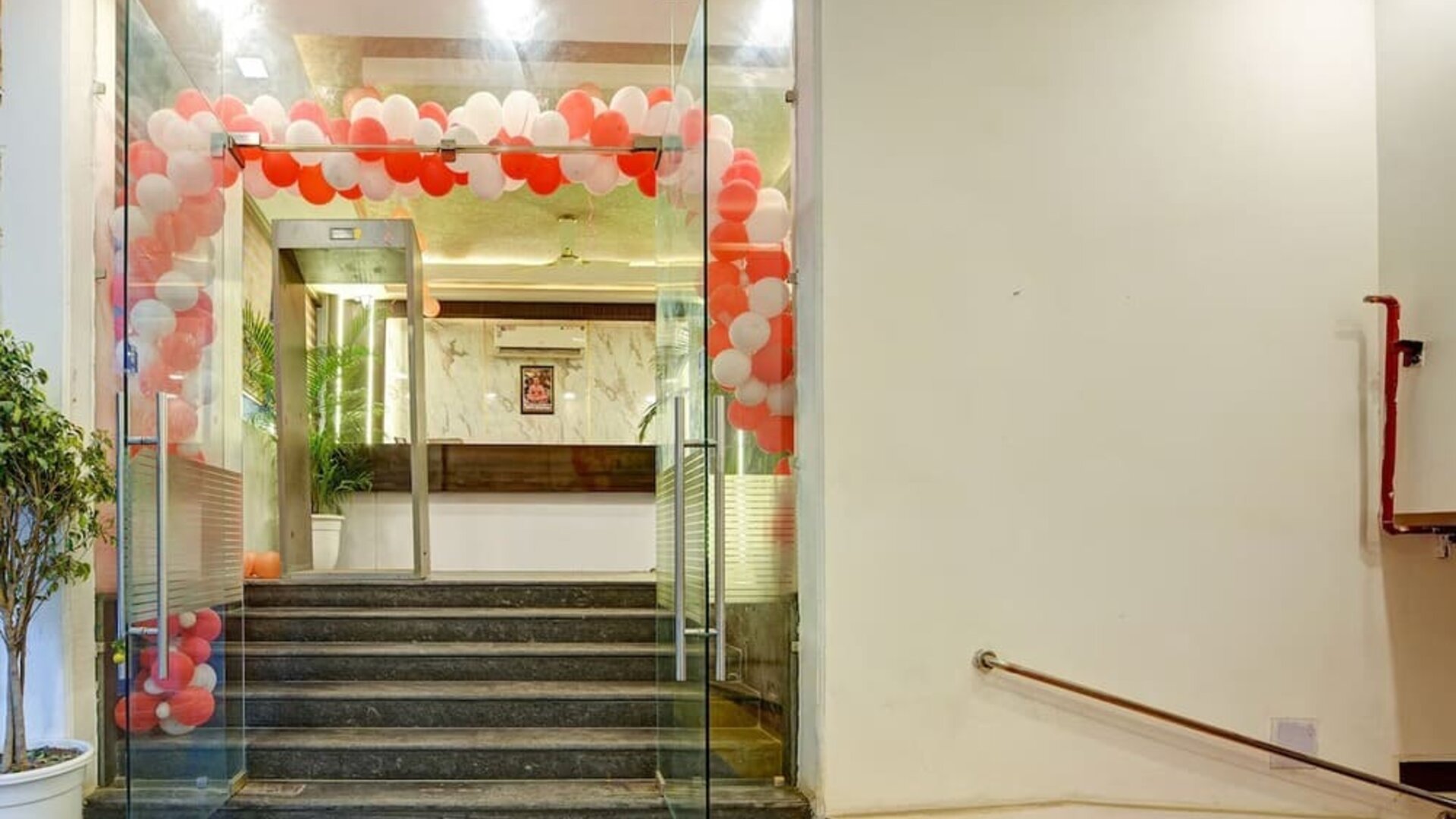 Glass entrance doors leading up several steps to the hotel reception area, decorated with an arch of red and white balloons, with a ramp and handrail visible to the right.