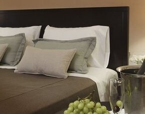 Close view of a king bed with layered pillows, dark headboard, bedside lamps, and champagne at El Conquistador Hotel.