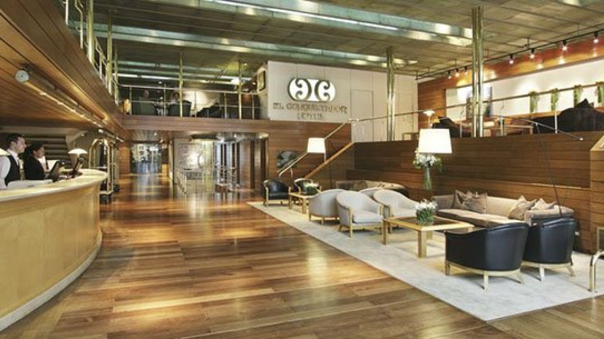 Spacious hotel lobby with a curved reception desk, wood flooring, lounge seating, and mezzanine at El Conquistador Hotel.
