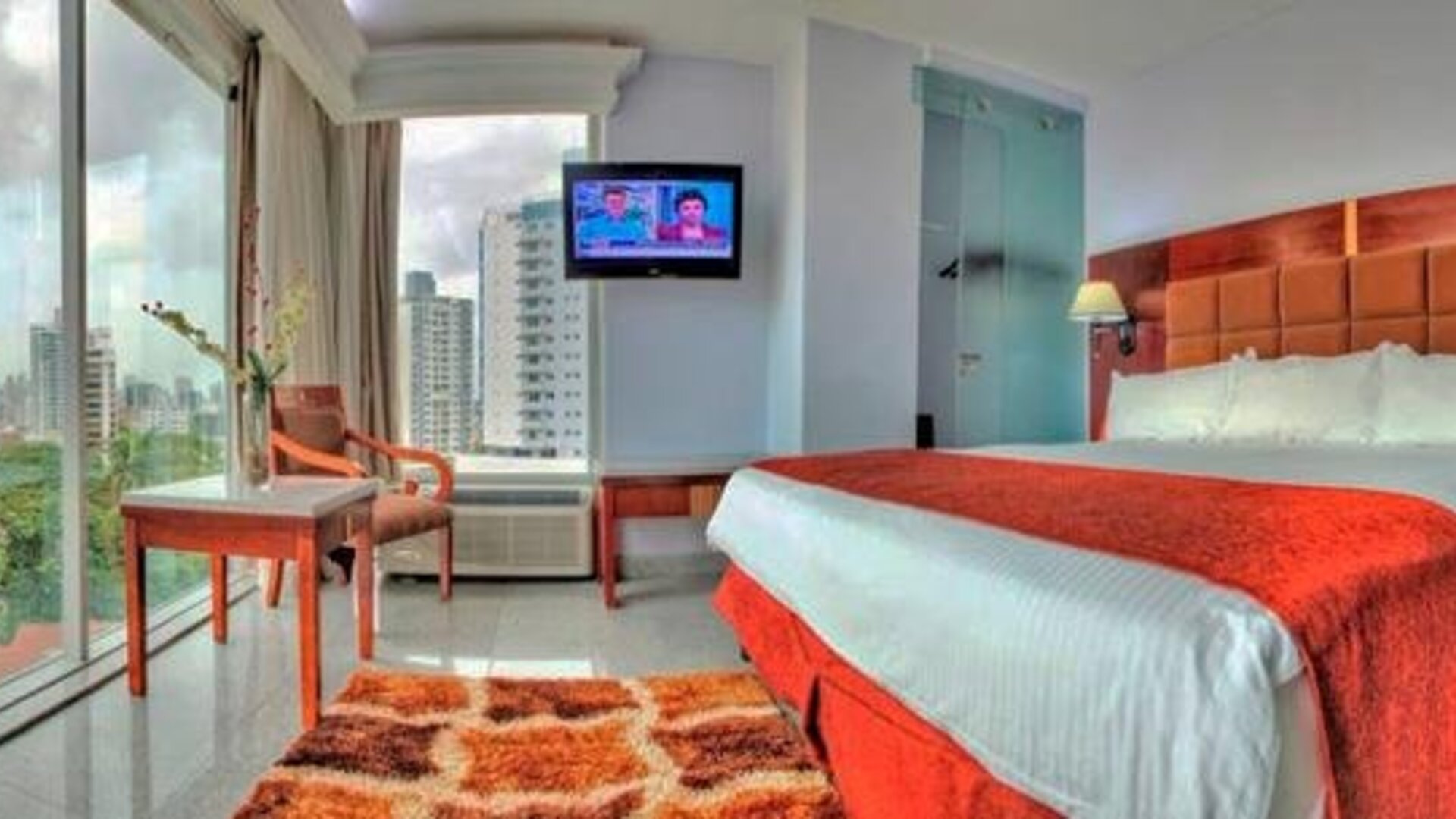 Guest room with a king bed, red runner, television, desk, chair, and floor-to-ceiling windows with city views at Hotel Grand International.