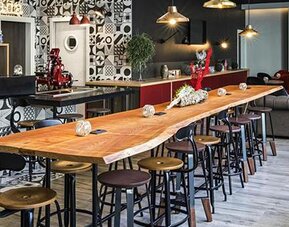 Hotel lounge and bar with a long wooden communal table, stools, sofas, pendant lights, and patterned walls at Hotel ibis Styles Barcelona City Bogatell.