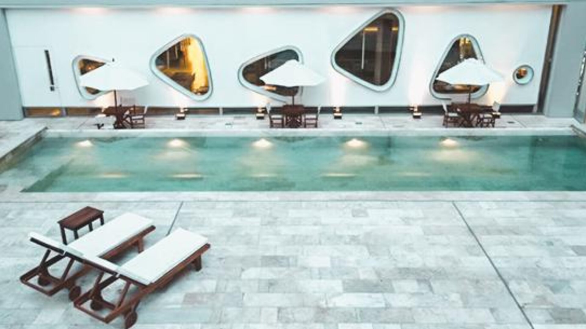 Indoor swimming pool with tiled deck, loungers, tables, umbrellas, and curved windows at Dazzler by Wyndham Buenos Aires Recoleta.
