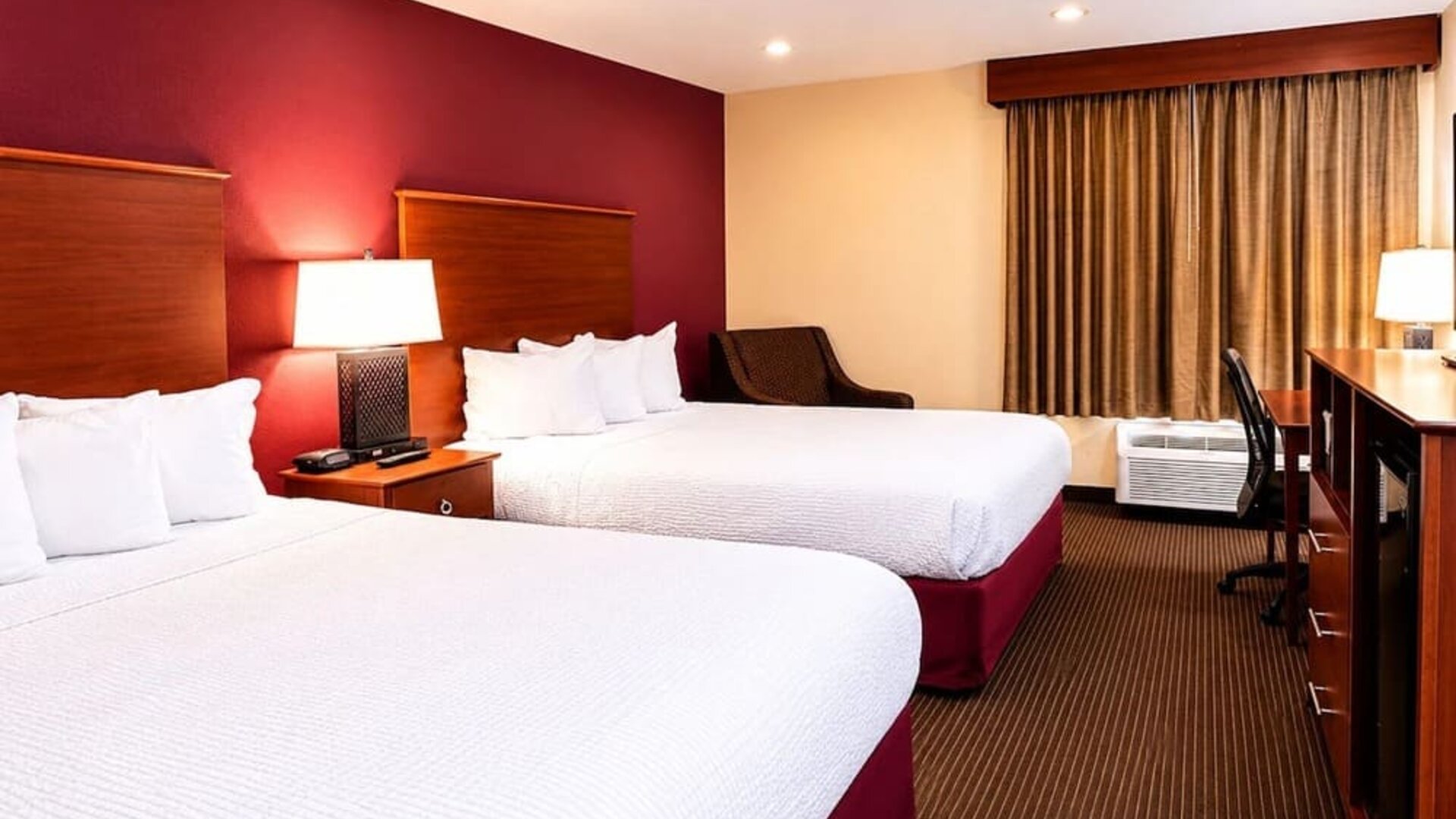 Spacious hotel guest room with two beds dressed in crisp white linens, rich red accent wall, warm wood headboards, bedside lamp, upholstered chair, work desk, air-conditioning unit and full-length curtains.