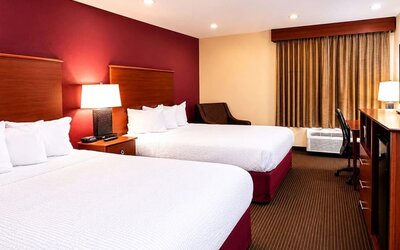 Spacious hotel guest room with two beds dressed in crisp white linens, rich red accent wall, warm wood headboards, bedside lamp, upholstered chair, work desk, air-conditioning unit and full-length curtains.