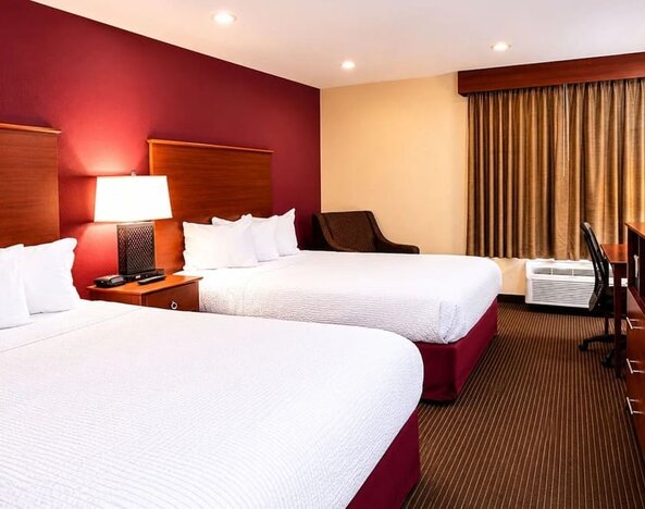 Spacious hotel guest room with two beds dressed in crisp white linens, rich red accent wall, warm wood headboards, bedside lamp, upholstered chair, work desk, air-conditioning unit and full-length curtains.