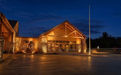 Lodge-style hotel exterior illuminated at night, featuring timber and stone architectural details, a peaked roof, large windows and outdoor seating near the entrance.