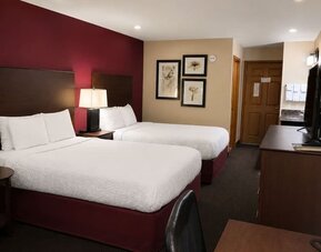 Comfortable hotel guest room with two beds dressed in white linens, burgundy accent wall and bed bases, dark wood headboards, bedside lamp, framed botanical artwork, television and wooden furnishings.