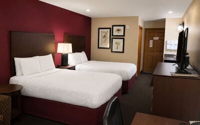Comfortable hotel guest room with two beds dressed in white linens, burgundy accent wall and bed bases, dark wood headboards, bedside lamp, framed botanical artwork, television and wooden furnishings.