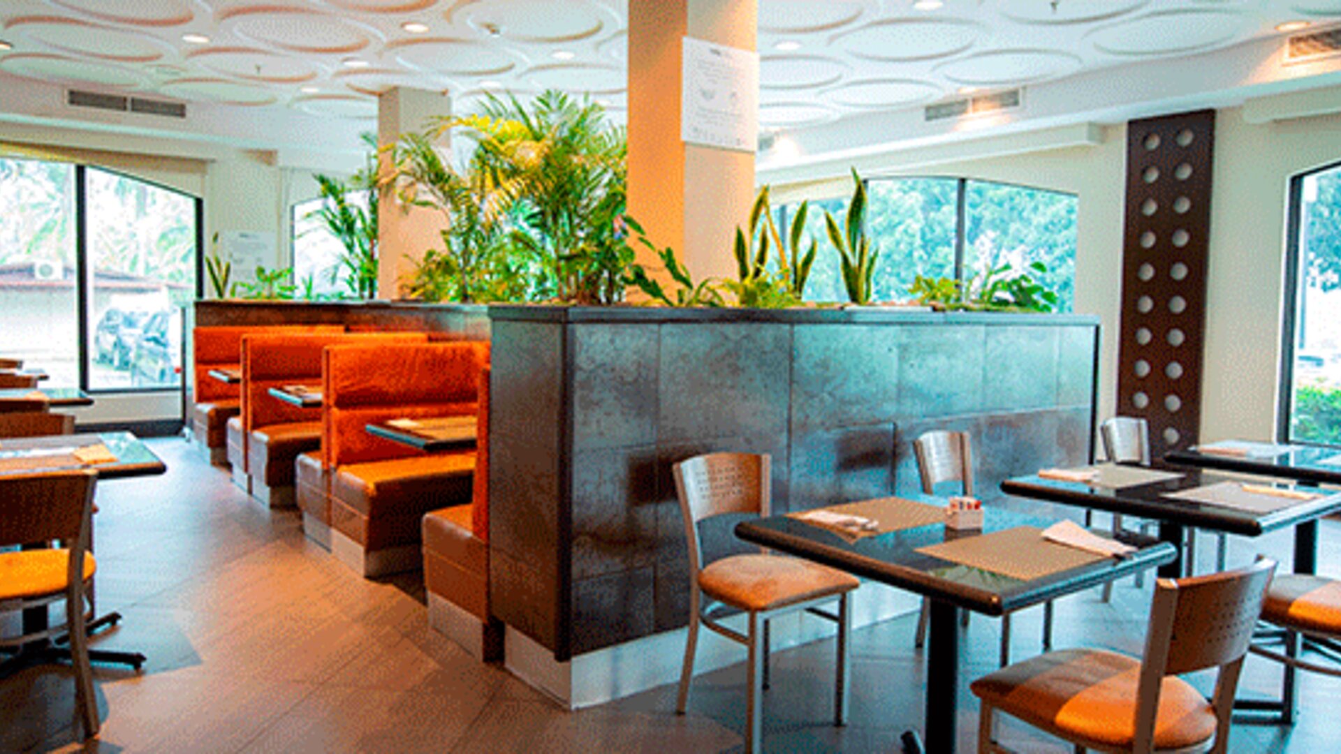 Hotel dining area with tables, chairs, orange booth seating and tropical plants beside large windows at Holiday Inn Panama Canal.
