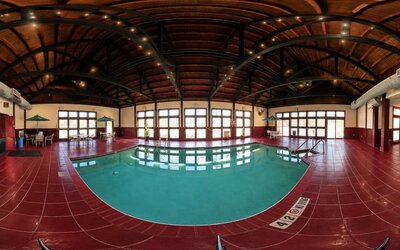 Spacious indoor hotel swimming pool surrounded by deep red tile flooring beneath a high, curved wood-paneled ceiling supported by dark exposed beams. Large windows and glass doors fill the pool area with natural light, while small seating areas with table