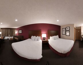 Comfortable hotel guest room featuring two beds with crisp white bedding and dark wooden headboards against a burgundy accent wall. The room includes a flat-screen TV, desk and chair, small seating area, bedside table with lamps, and curtained window. A v