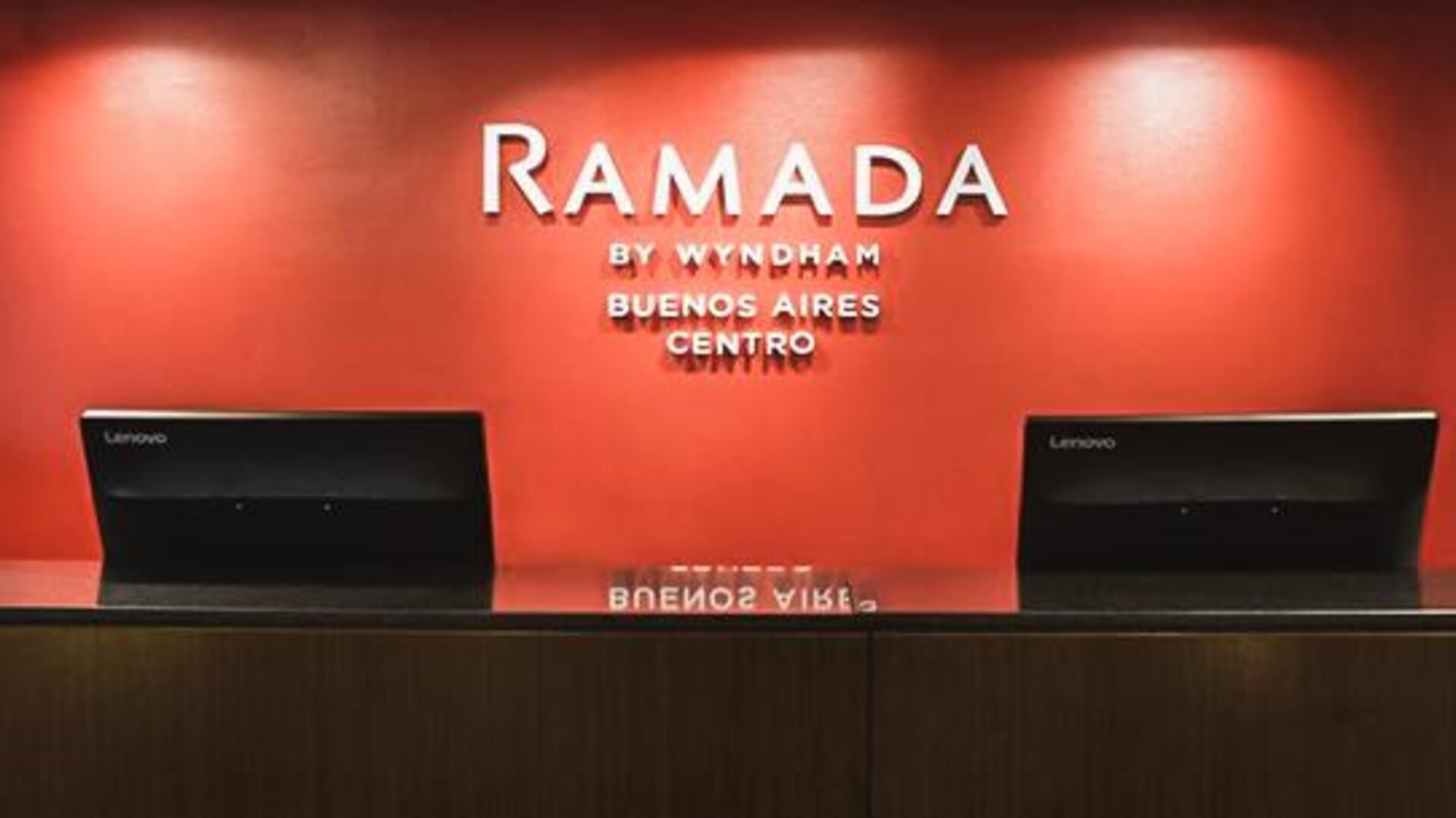 Hotel reception desk with two computer terminals and a red accent wall displaying the Ramada by Wyndham Buenos Aires Centro logo.