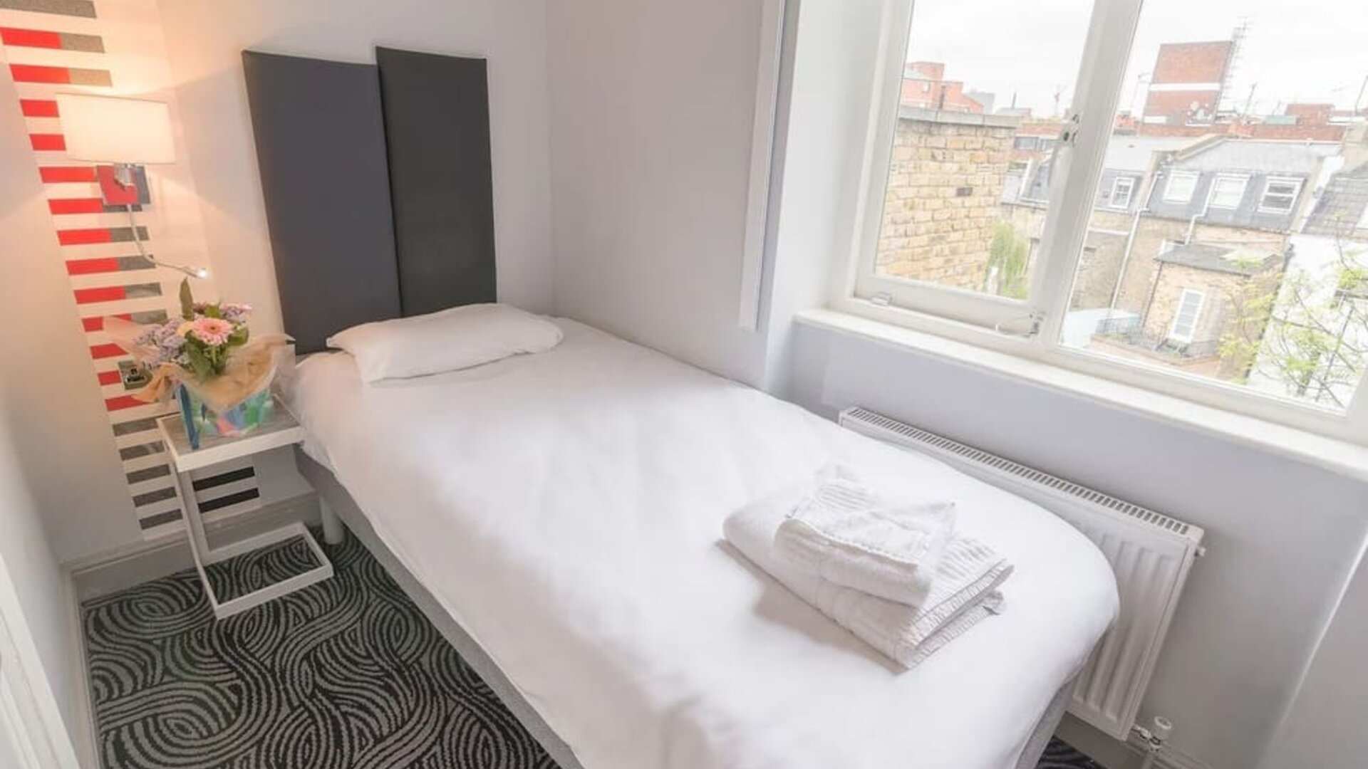 Single guest room with a white bed, dark upholstered headboard, bedside table, flowers and large window overlooking surrounding buildings at Victoria Inn.