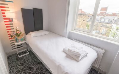 Single guest room with a white bed, dark upholstered headboard, bedside table, flowers and large window overlooking surrounding buildings at Victoria Inn.