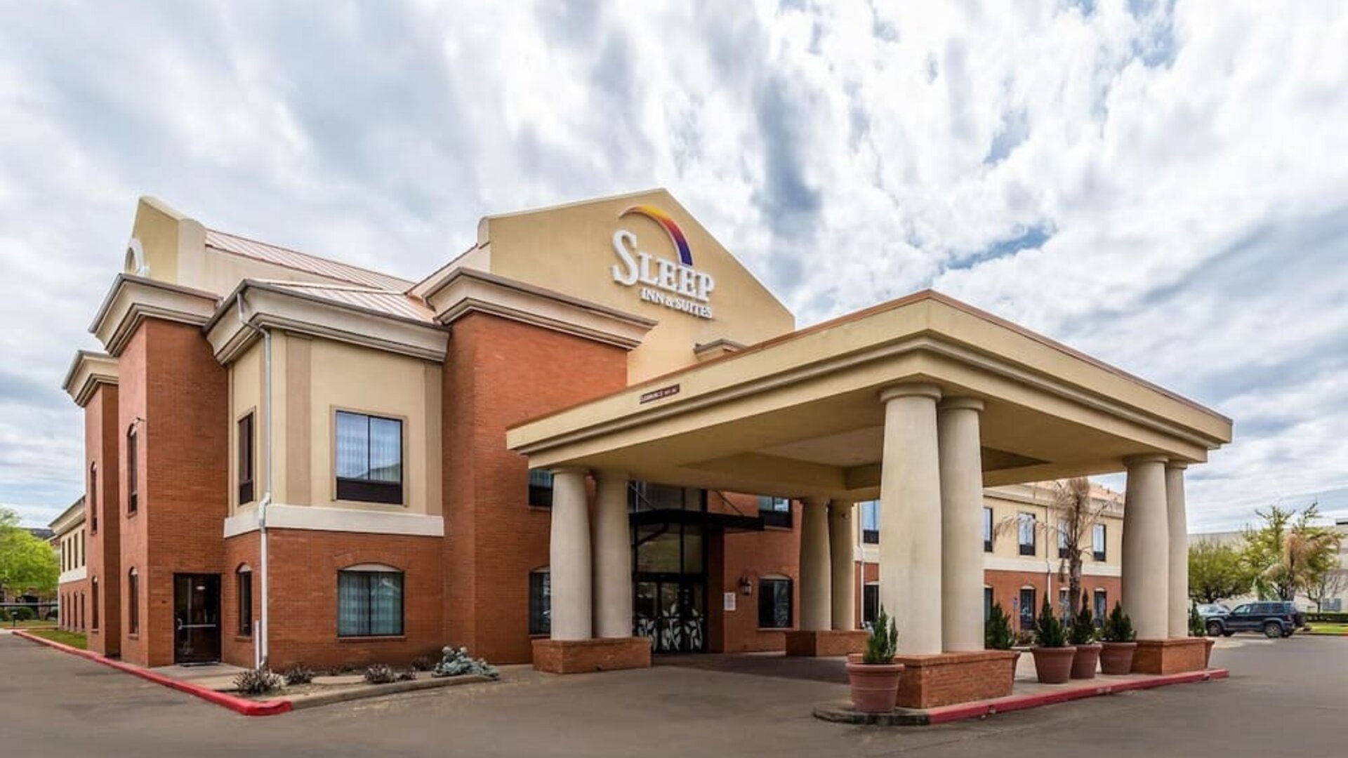 Exterior of Sleep Inn & Suites Stafford - Sugar Land, showing the brick-and-beige hotel building, covered entrance with columns, parking area, and landscaped planters.