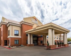Exterior of Sleep Inn & Suites Stafford - Sugar Land, showing the brick-and-beige hotel building, covered entrance with columns, parking area, and landscaped planters.
