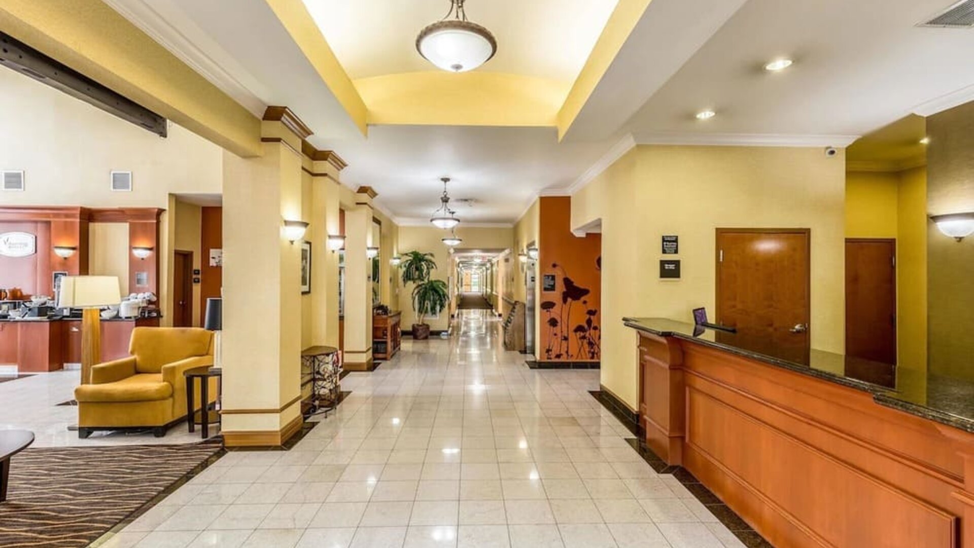 Hotel reception area with a long wooden front desk, yellow walls, seating area, tiled floor, and hallway leading to guest rooms at Sleep Inn & Suites Stafford - Sugar Land.