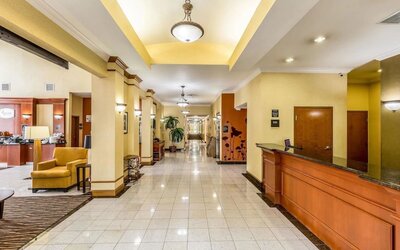 Hotel reception area with a long wooden front desk, yellow walls, seating area, tiled floor, and hallway leading to guest rooms at Sleep Inn & Suites Stafford - Sugar Land.