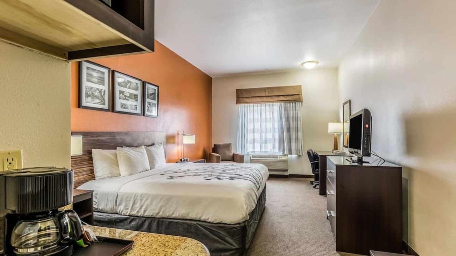 Spacious king guest room with a large bed, orange accent wall, framed artwork, sofa, ottoman, armchair, television, kitchenette, refrigerator, and coffee maker at Sleep Inn & Suites Stafford - Sugar Land.