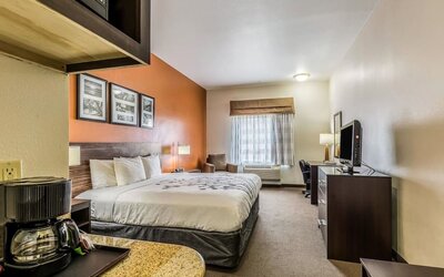 Spacious king guest room with a large bed, orange accent wall, framed artwork, sofa, ottoman, armchair, television, kitchenette, refrigerator, and coffee maker at Sleep Inn & Suites Stafford - Sugar Land.