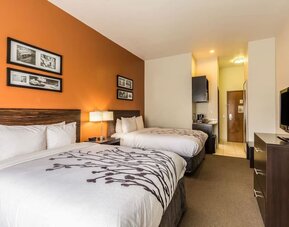Double guest room with two beds, white bedding, orange accent wall, framed artwork, bedside lamp, television, desk, refrigerator, microwave, and coffee maker at Sleep Inn & Suites Stafford - Sugar Land.