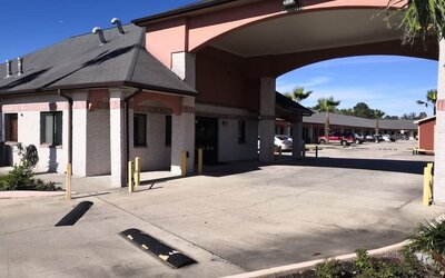 Exterior of Scottish Inn & Suites showing the low-rise hotel buildings, covered entrance with large arches, parking area, palm trees, and landscaped grounds.